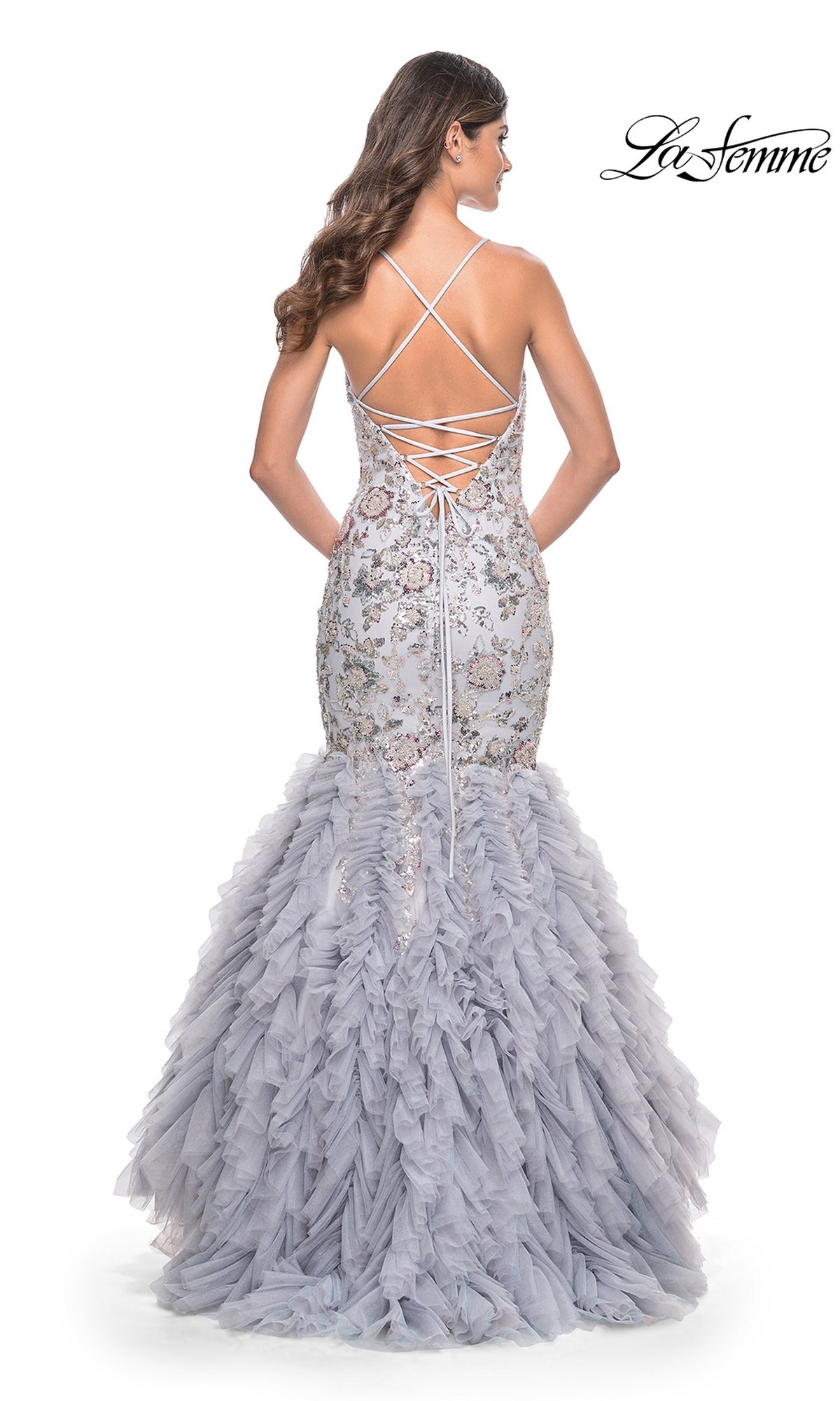 A woman models the La Femme Silver Ruffled Long Prom Dress 32105—a backless mermaid gown in light gray with lace-up details and a ruffled tulle skirt, beautifully displayed from the back.